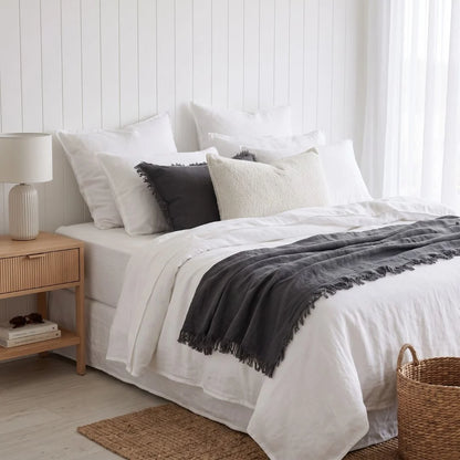 A neatly made bed in a bright bedroom setting, dressed in white bedding with a dark charcoal throw blanket draped across the foot. The bed is styled with multiple white and dark gray pillows, a light wood nightstand with a white lamp on the left, and a woven basket on the floor at the right.