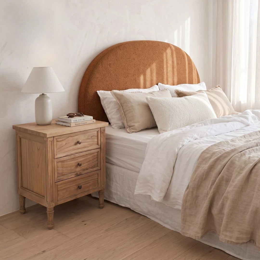 A styled bedroom scene featuring a light oak nightstand with a white table lamp, next to a bed with a large, curved, warm brown upholstered headboard, white linens, and beige accents.