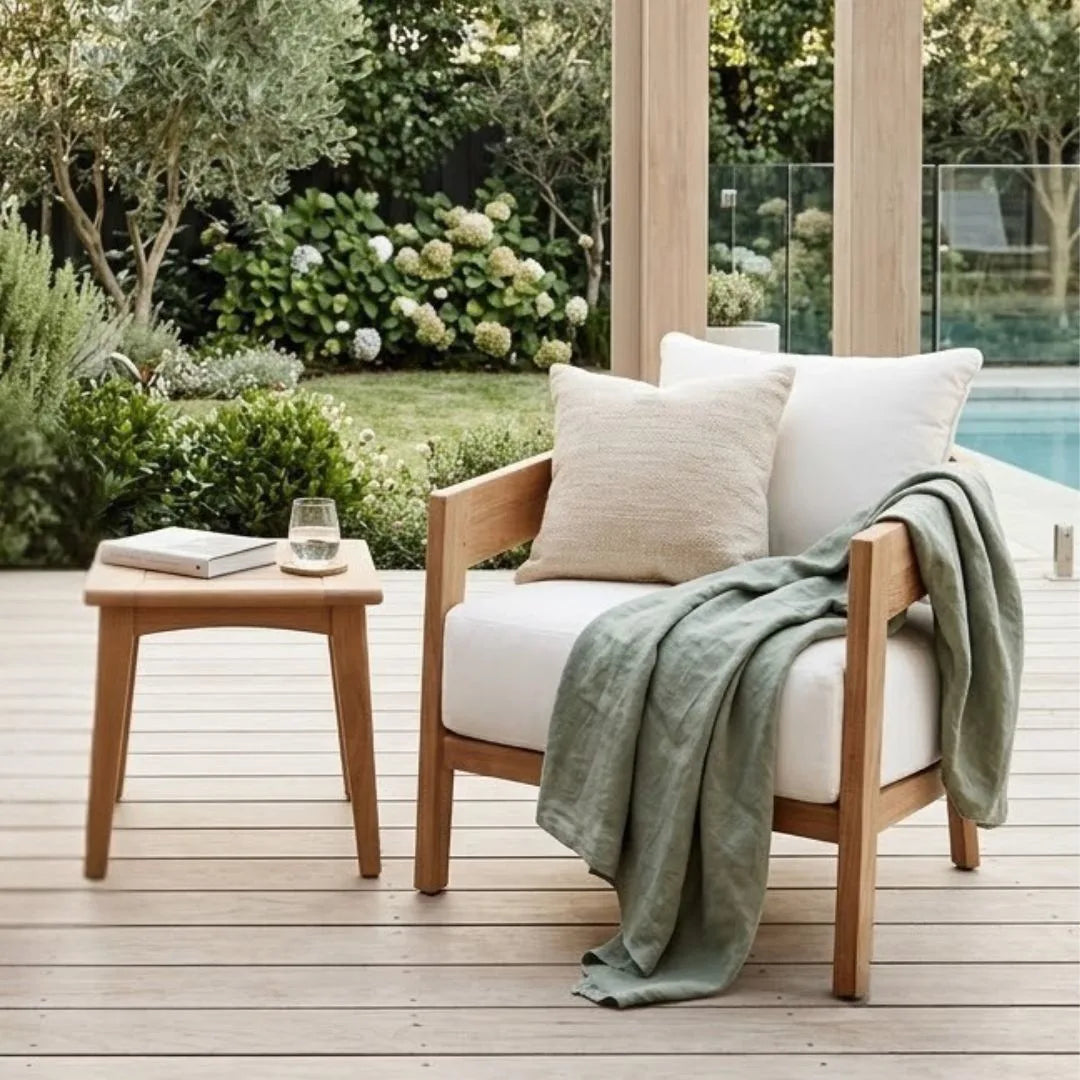 Wooden chair with white cushions and a green blanket on a wooden deck with a garden view.