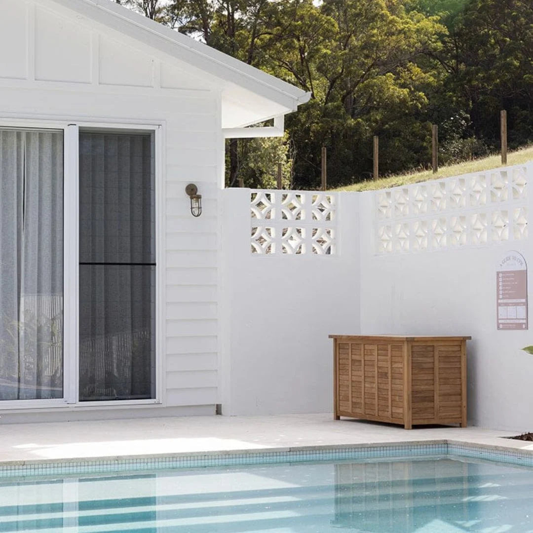 A wooden outdoor storage cabinet with a slatted front and closed doors, placed on a patio beside a swimming pool and white house walls.