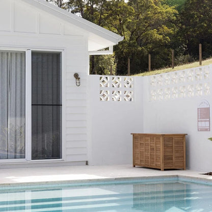A wooden outdoor storage cabinet with a slatted front and closed doors, placed on a patio beside a swimming pool and white house walls.