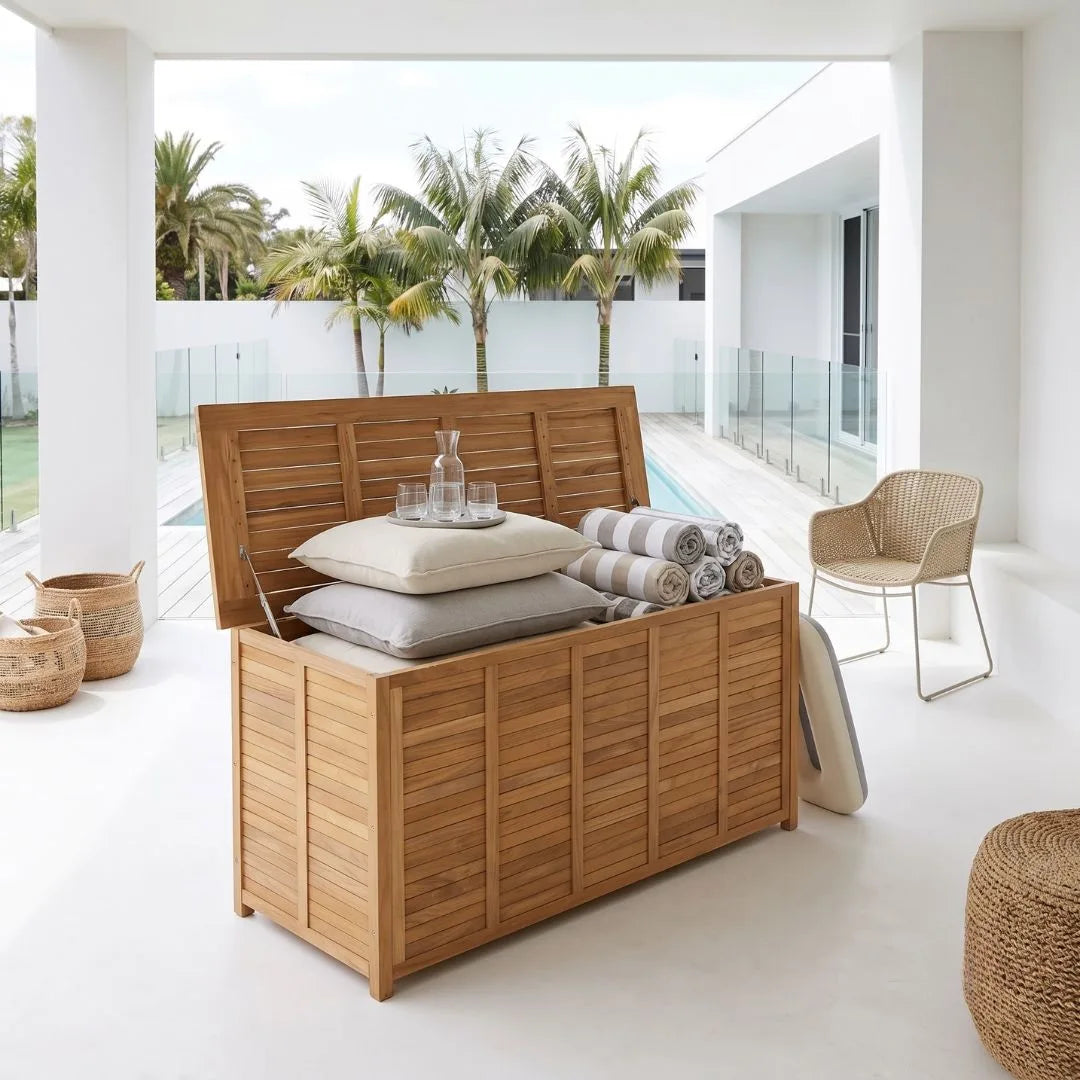 A large wooden storage trunk with a slatted lid sits open in a bright modern patio setting, styled with pillows, rolled towels, and a glass decanter. A woven chair, baskets, and palm trees are visible in the background.