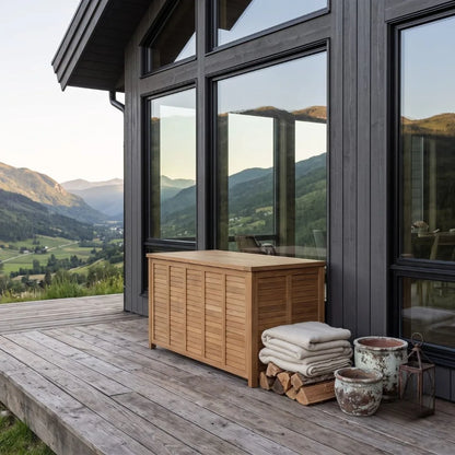 A wooden outdoor storage chest with a slatted front sits on a wooden deck beside a modern house with large windows overlooking a mountain valley.