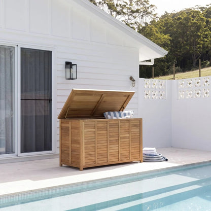 A wooden outdoor storage chest with a lift-up lid sits on a patio beside a swimming pool. The chest has a warm medium-brown wood finish and is shown open with a striped cushion inside, set against a white house and green trees in the background.