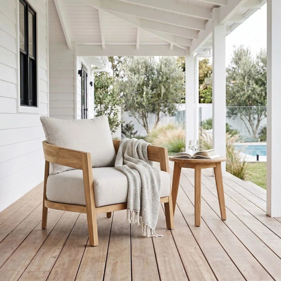A light wood outdoor armchair with off-white cushions and a draped knit throw blanket, placed on a wooden porch beside a small round wooden side table with an open book.