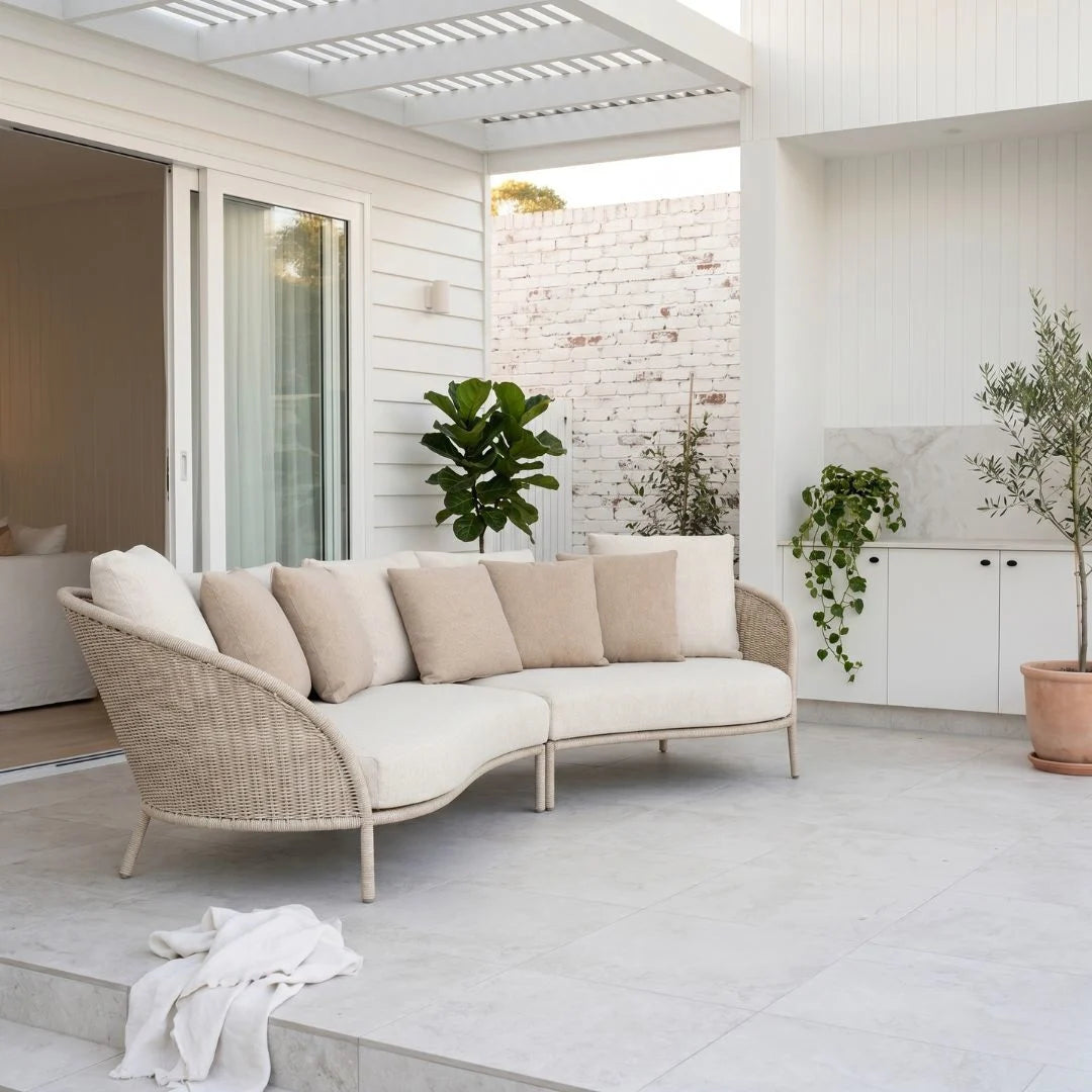 Modern outdoor seating area with a curved wicker sofa, plants, and a white wall.