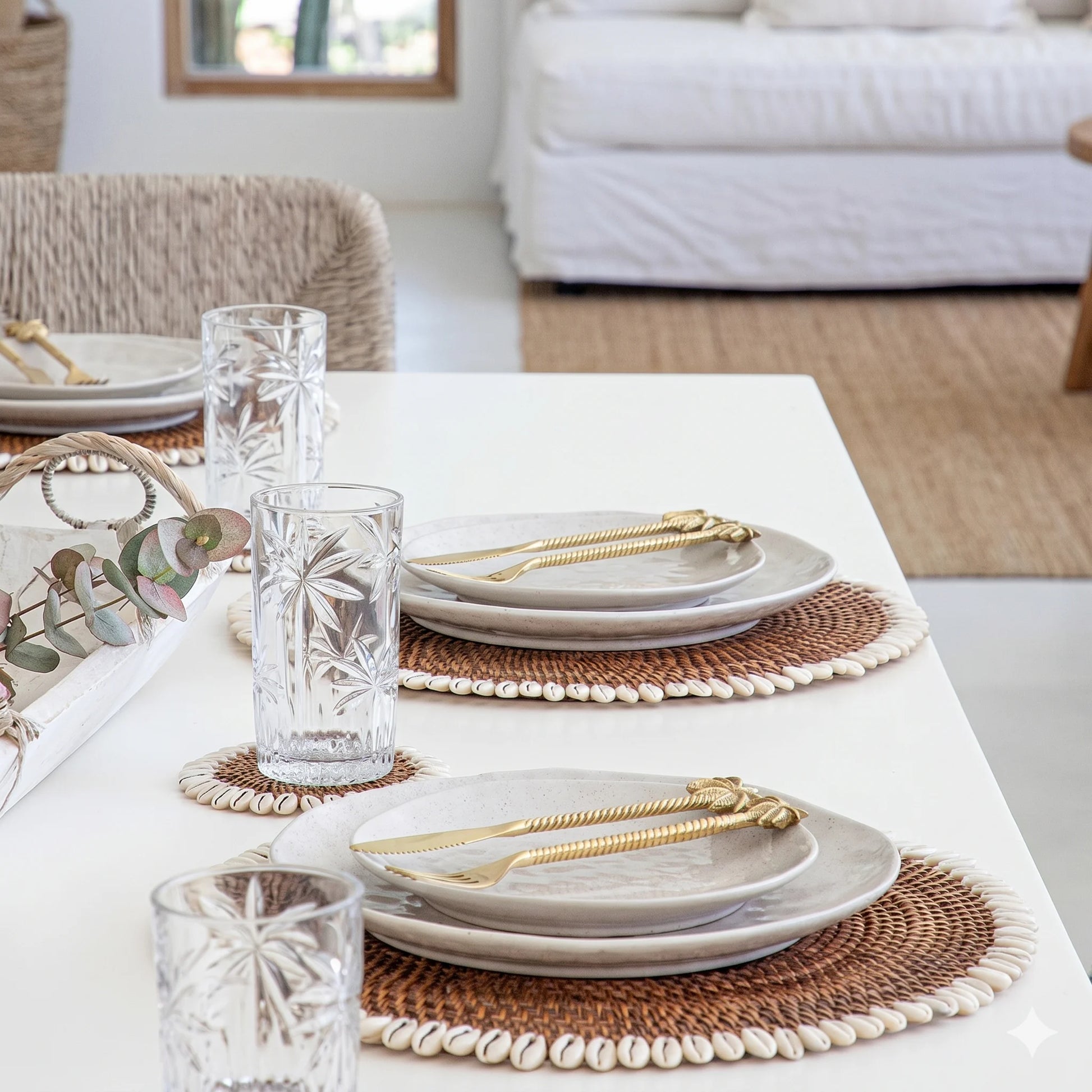 Dining table set with plates, glasses, and gold cutlery on woven placemats.
