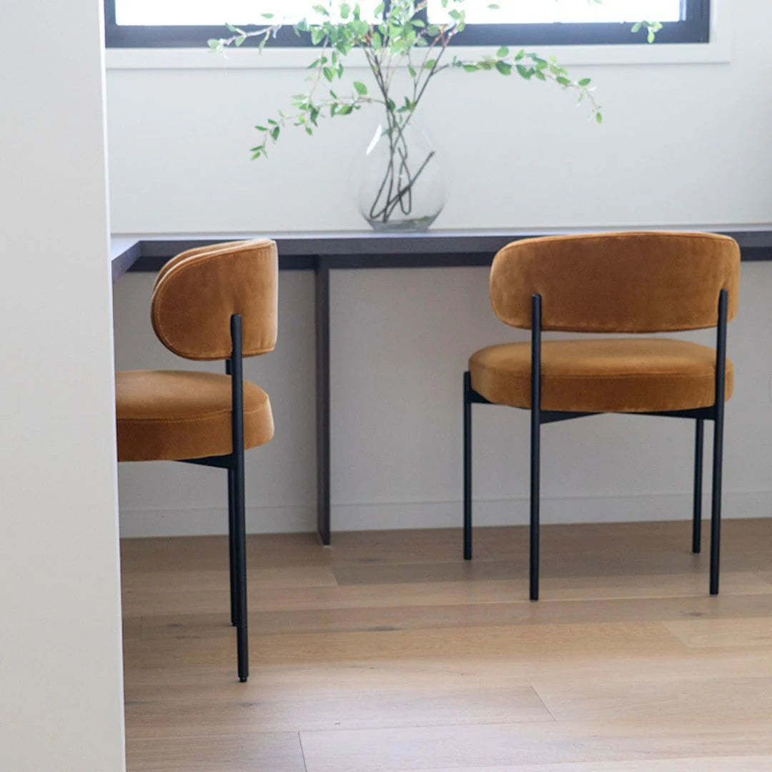 Two brown chairs at a dining table with a vase of greenery on a white wall.