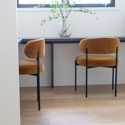 Two brown chairs at a dining table with a vase of greenery on a white wall.