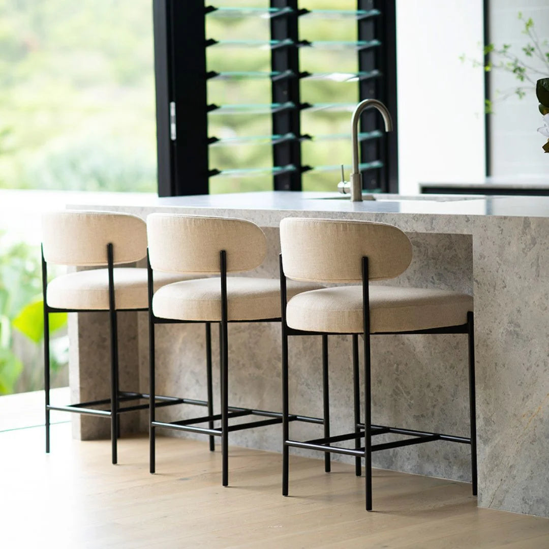 Three beige cushioned bar stools with black metal legs in a modern kitchen setting.