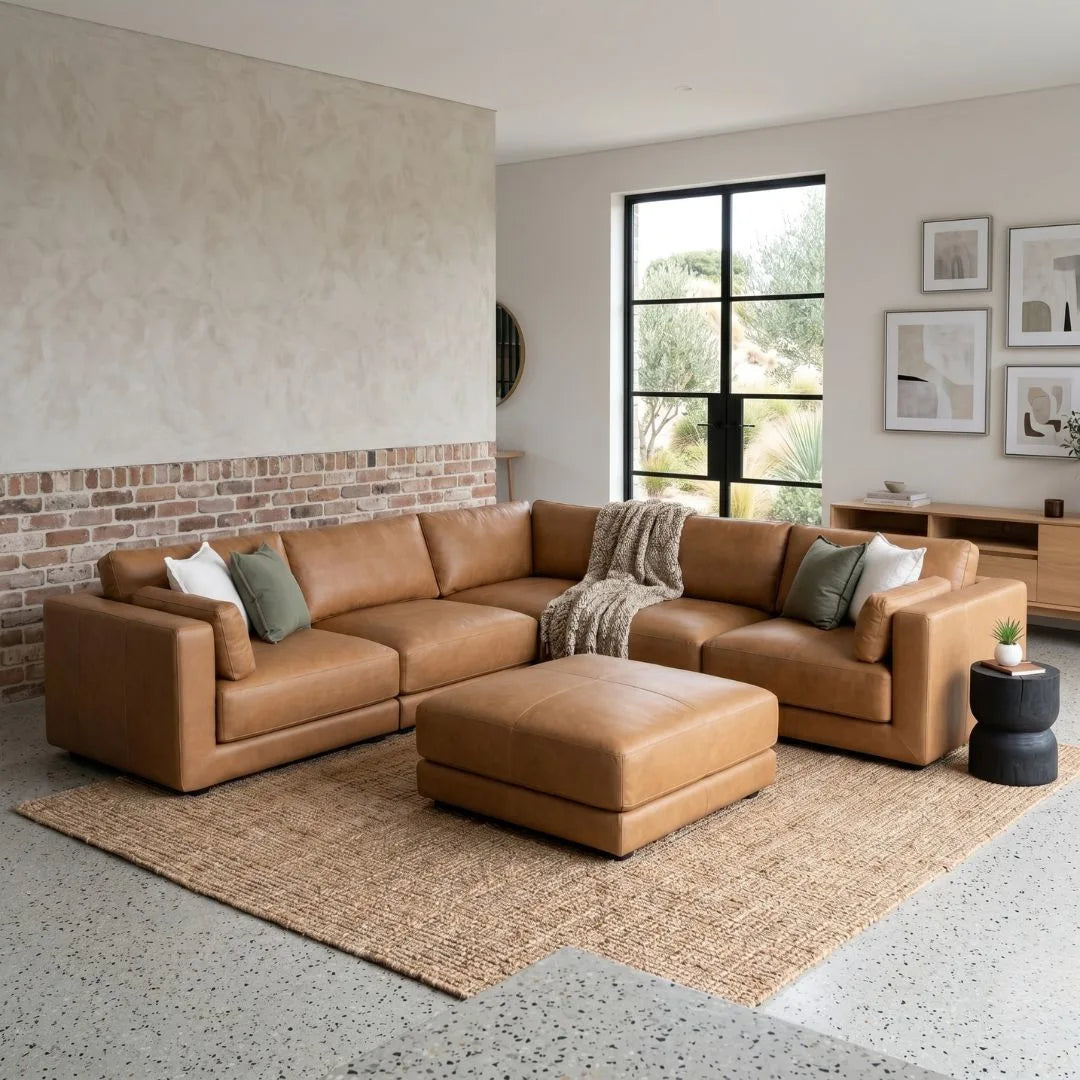 Brown leather sectional sofa in a modern living room with a brick wall and large window.