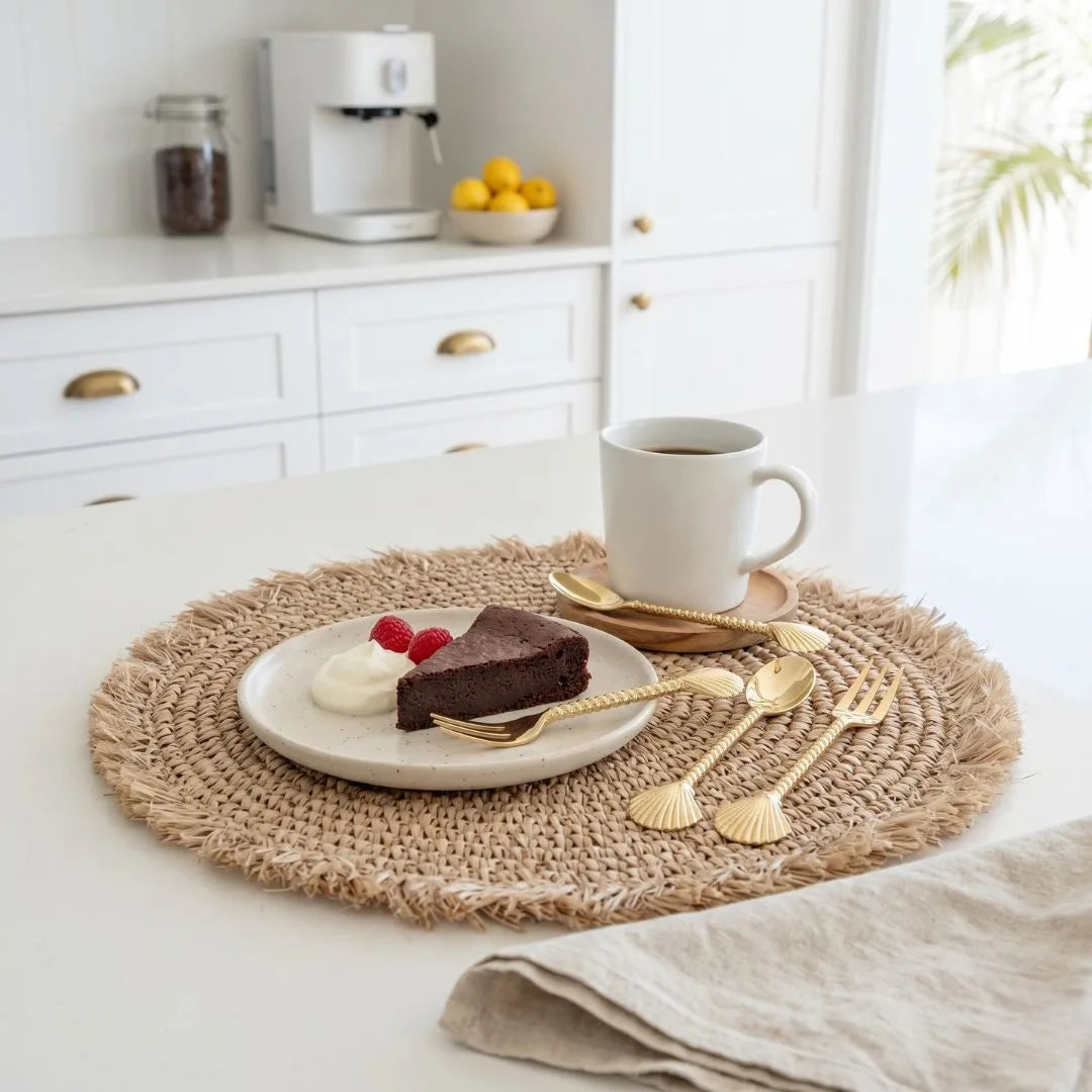 Cafeteria setting with a plate of cake, a cup of coffee, and gold cutlery on a woven placemat.
