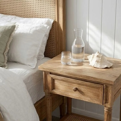 Wooden nightstand with a glass carafe and shell next to a bed with white bedding.