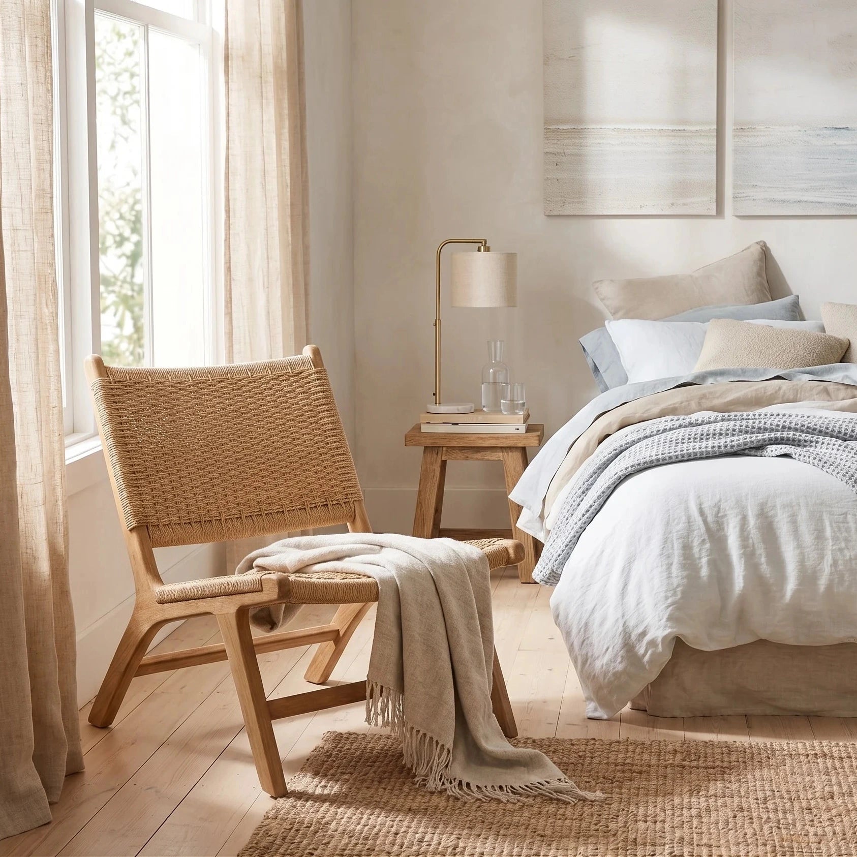 A styled bedroom scene with a bed dressed in neutral-toned bedding and a textured throw blanket, beside a woven wood chair with a draped throw. A small side table with a lamp sits between the chair and the bed, and wall art hangs above the headboard.