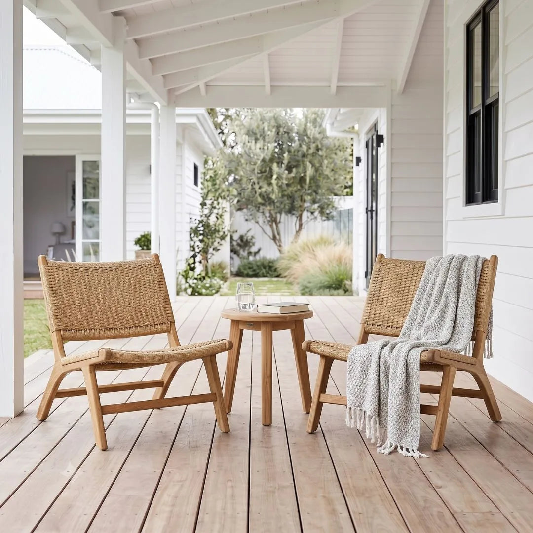 Two woven rattan lounge chairs with light wood frames on a wooden porch, with a small round wooden side table between them and a white throw blanket draped over one chair. A house exterior and garden are visible in the background.