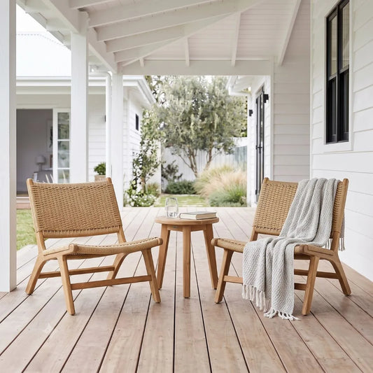 Two woven rattan lounge chairs with light wood frames on a wooden porch, with a small round wooden side table between them and a white throw blanket draped over one chair. A house exterior and garden are visible in the background.