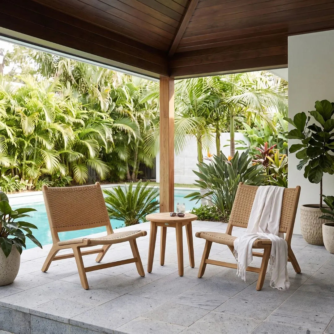 Two wooden lounge chairs with woven seats and a small round wooden side table on a stone patio, with a white throw draped over one chair. The setting is outdoors under a covered wooden roof with lush tropical plants and a swimming pool in the background.