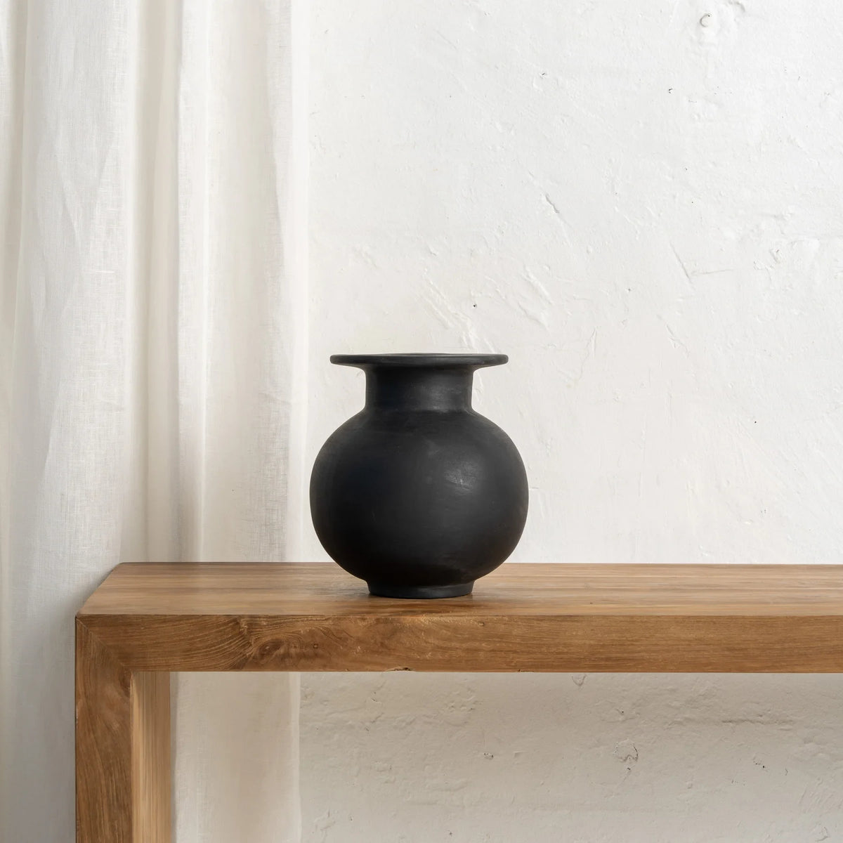 Black vase on a wooden table with a white curtain in the background