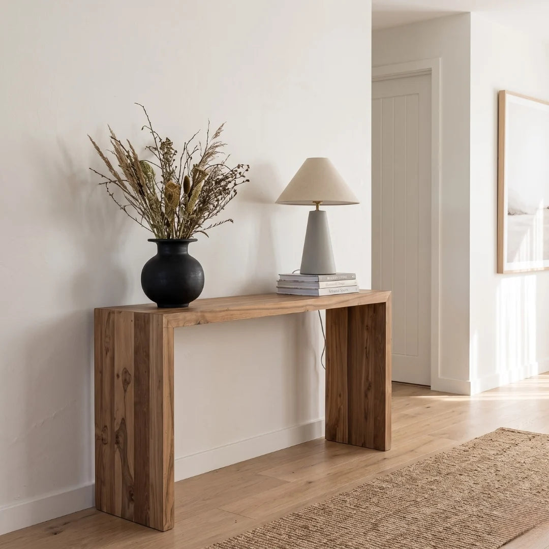 A rustic wooden console table in a warm medium brown finish, styled with a black vase of dried grasses and a gray table lamp in a bright, modern room with light wood floors and a beige rug.