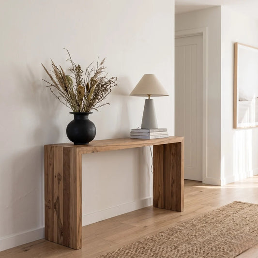A rustic wooden console table in a warm medium brown finish, styled with a black vase of dried grasses and a gray table lamp in a bright, modern room with light wood floors and a beige rug.