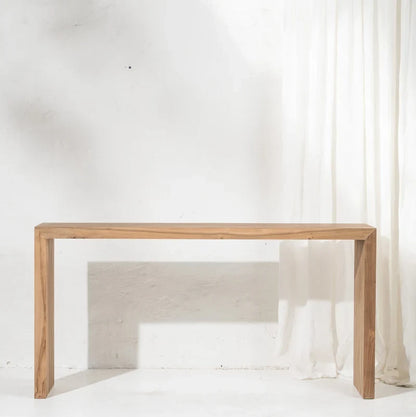 A minimalist light oak wood console table with a live-edge top and block legs, set against a white textured wall with a sheer white curtain on the right.