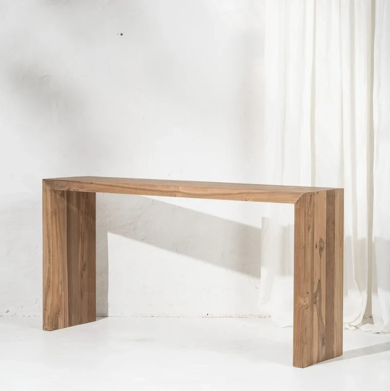 A minimalist rectangular wooden console table with a natural brown wood finish, shown against a white textured wall with a white curtain on the right.