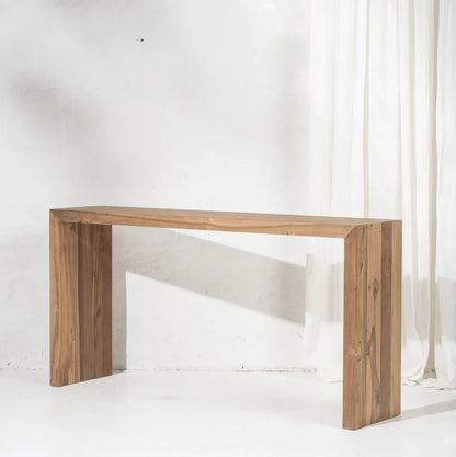 A minimalist rectangular wooden console table with a natural brown wood finish, shown against a white textured wall with a white curtain on the right.