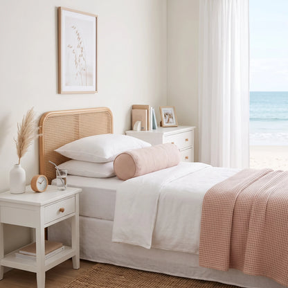 A neatly made bed in a bright, minimalist bedroom with white linens, a blush pink cylindrical pillow, a woven cane headboard, and a window looking out to the ocean.
