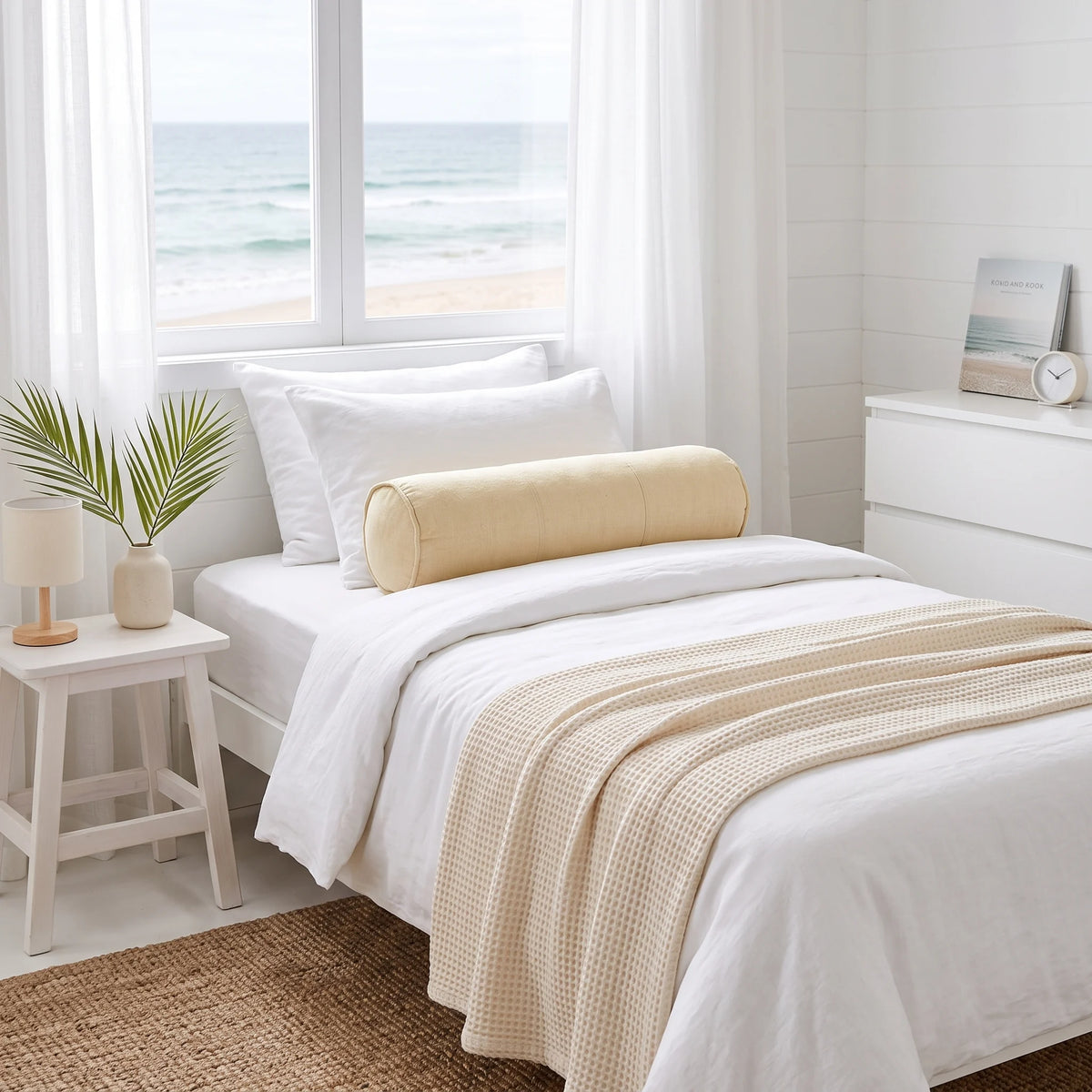 A neatly made bed in a bright, minimalist bedroom with a white duvet, white pillows, a beige cylindrical bolster pillow, and a beige waffle-knit throw blanket. A window behind the bed shows a view of the ocean, and a small side table with a plant is on the left.
