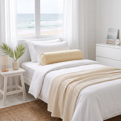 A neatly made bed in a bright, minimalist bedroom with a white duvet, white pillows, a beige cylindrical bolster pillow, and a beige waffle-knit throw blanket. A window behind the bed shows a view of the ocean, and a small side table with a plant is on the left.
