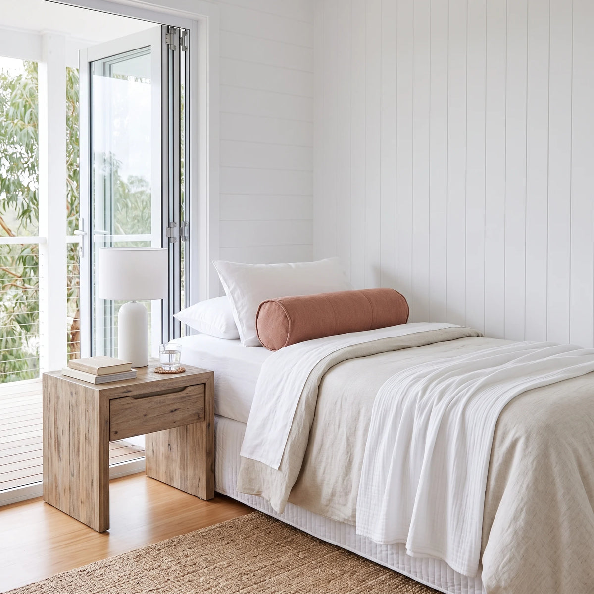 A minimalist single bed styled with white linens, a dusty rose cylindrical bolster pillow, and a white table lamp on a light wood nightstand in a bright, modern bedroom.