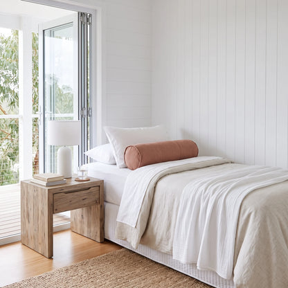 A minimalist single bed styled with white linens, a dusty rose cylindrical bolster pillow, and a white table lamp on a light wood nightstand in a bright, modern bedroom.