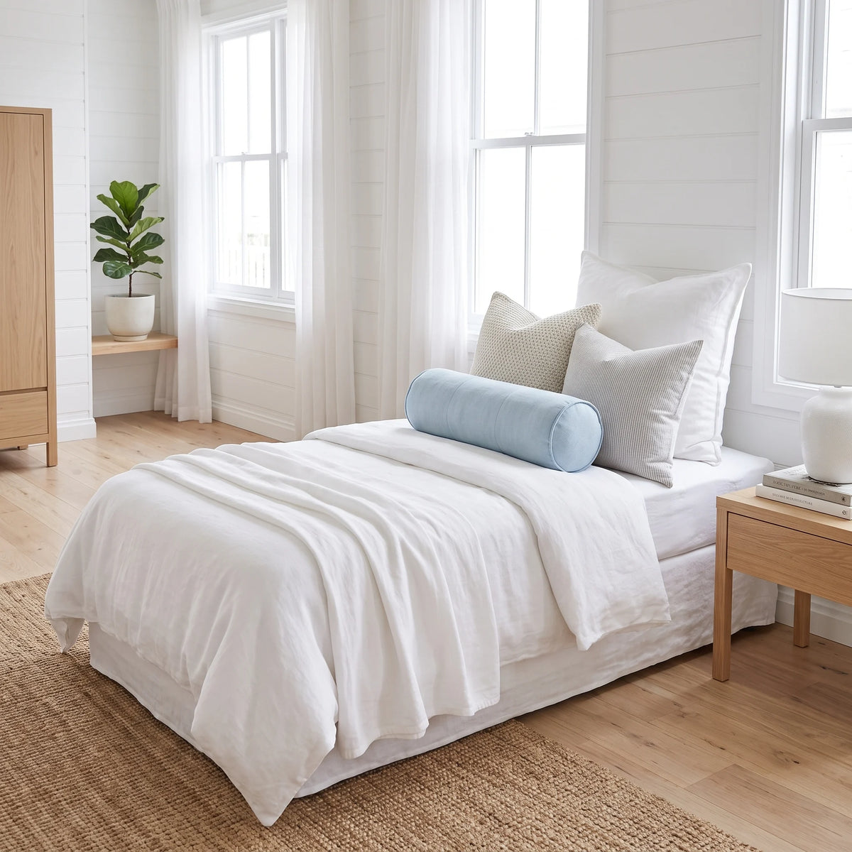 A neatly made bed in a bright, minimalist bedroom. The bedding set is predominantly white with a light blue cylindrical bolster pillow and a textured beige accent pillow.