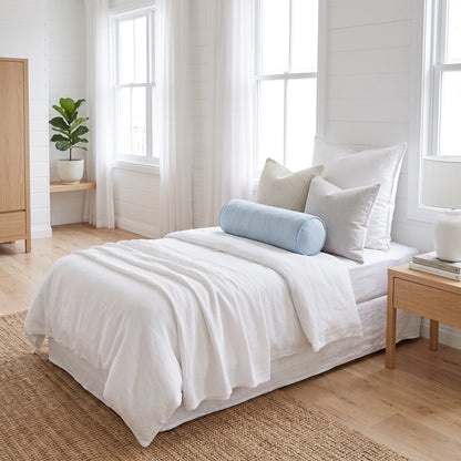 A neatly made bed in a bright, minimalist bedroom. The bedding set is predominantly white with a light blue cylindrical bolster pillow and a textured beige accent pillow.