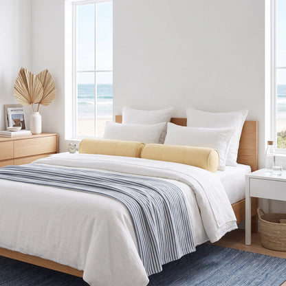 A neatly made bed in a bright beach-style bedroom, featuring white bedding, two pale yellow cylindrical bolster pillows, a blue-and-white striped throw blanket, a light wood bed frame, and large windows overlooking the ocean.