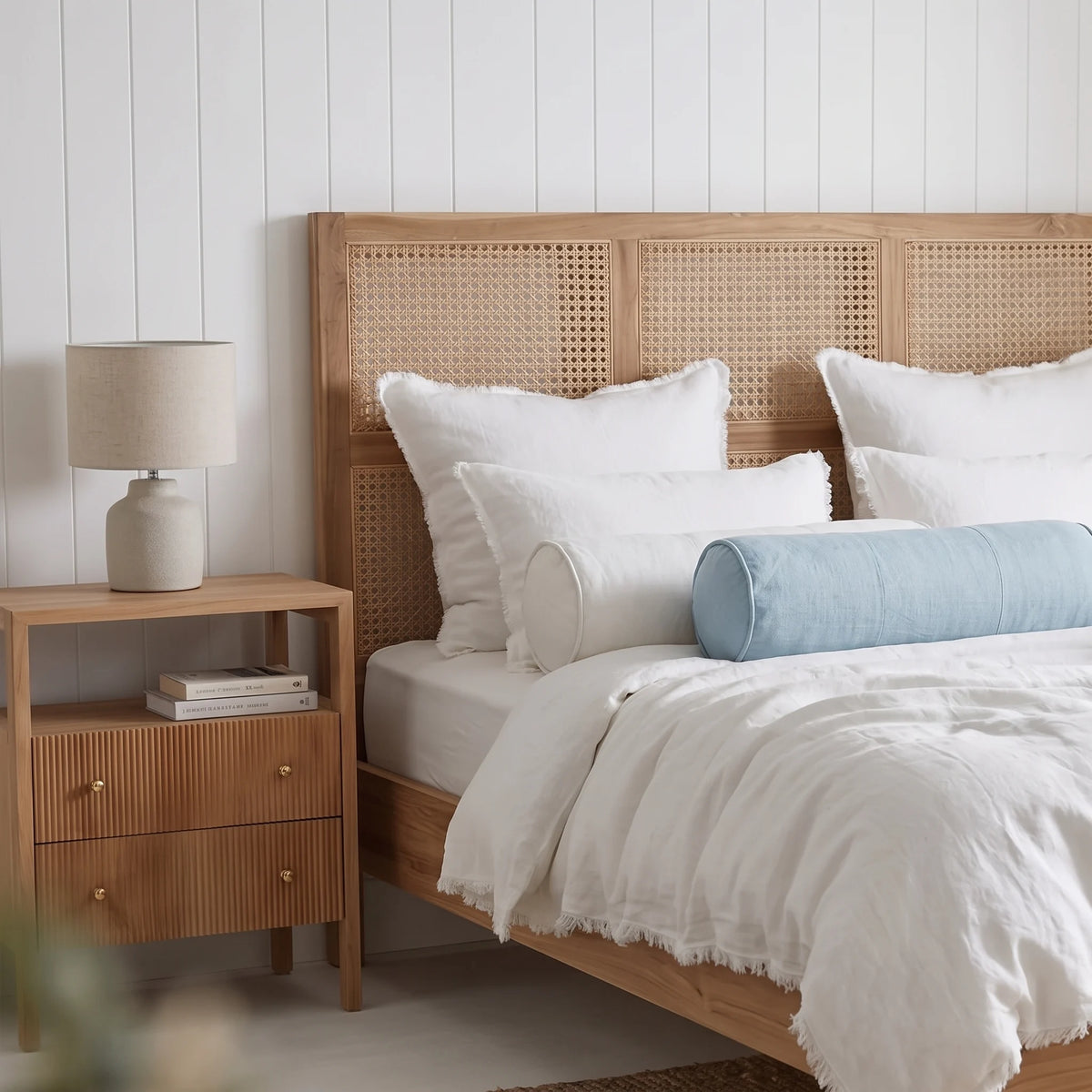 A wooden bed frame with a woven cane headboard, styled with white linens, pillows, and a light blue cylindrical bolster pillow, next to a matching wooden nightstand with a lamp.