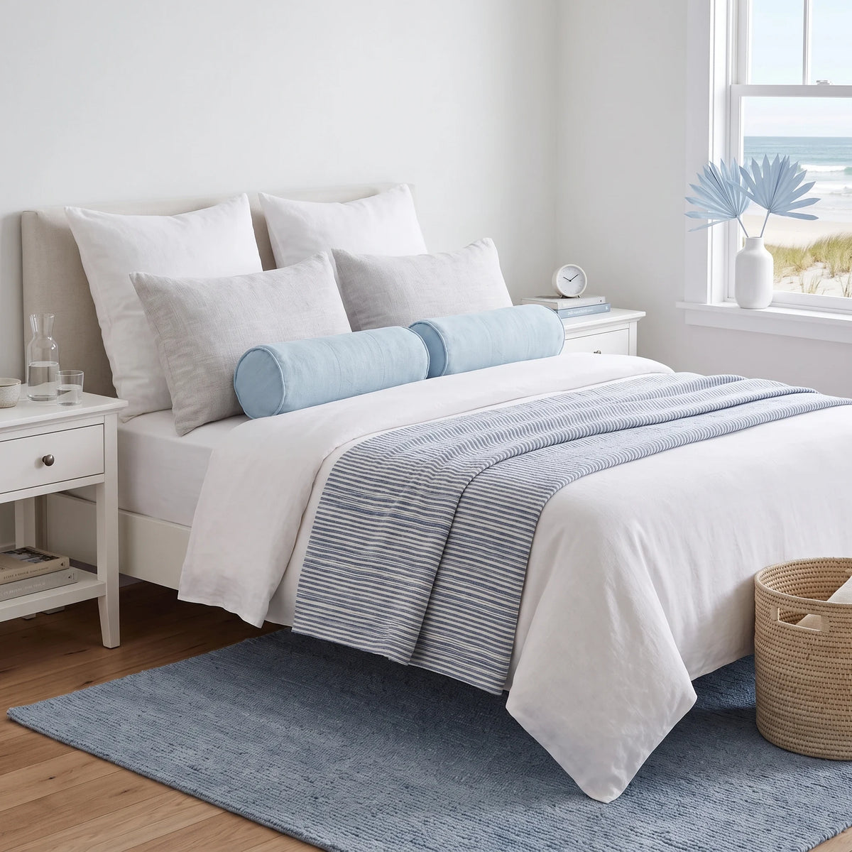 A neatly made bed in a bright bedroom setting, styled with white bedding, light blue cylindrical pillows, a blue-and-white striped throw blanket, a blue rug, and a woven basket on the floor.