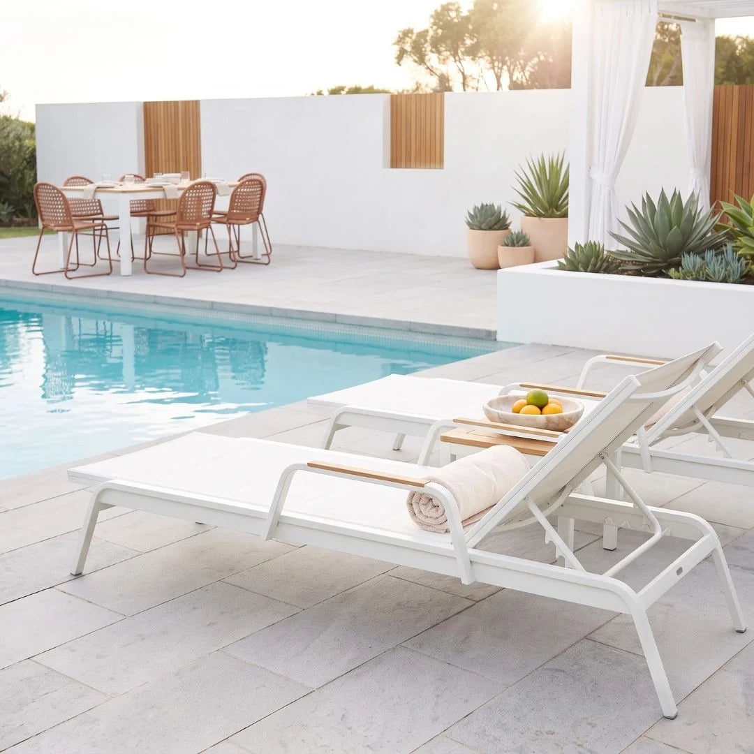 White outdoor patio chaise lounge chairs with light wood armrests arranged beside a swimming pool on a stone patio, with a dining set and plants in the background.
