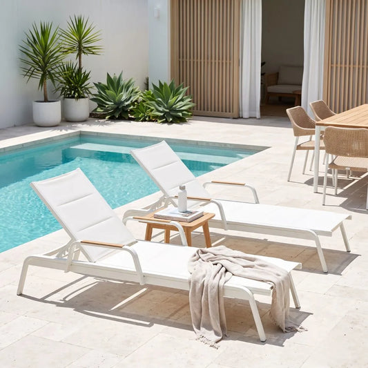 Two white outdoor chaise lounge chairs with light wood armrests are placed beside a swimming pool on a light stone patio. A small wooden side table sits between them, and a beige throw blanket is draped over the foreground chair.