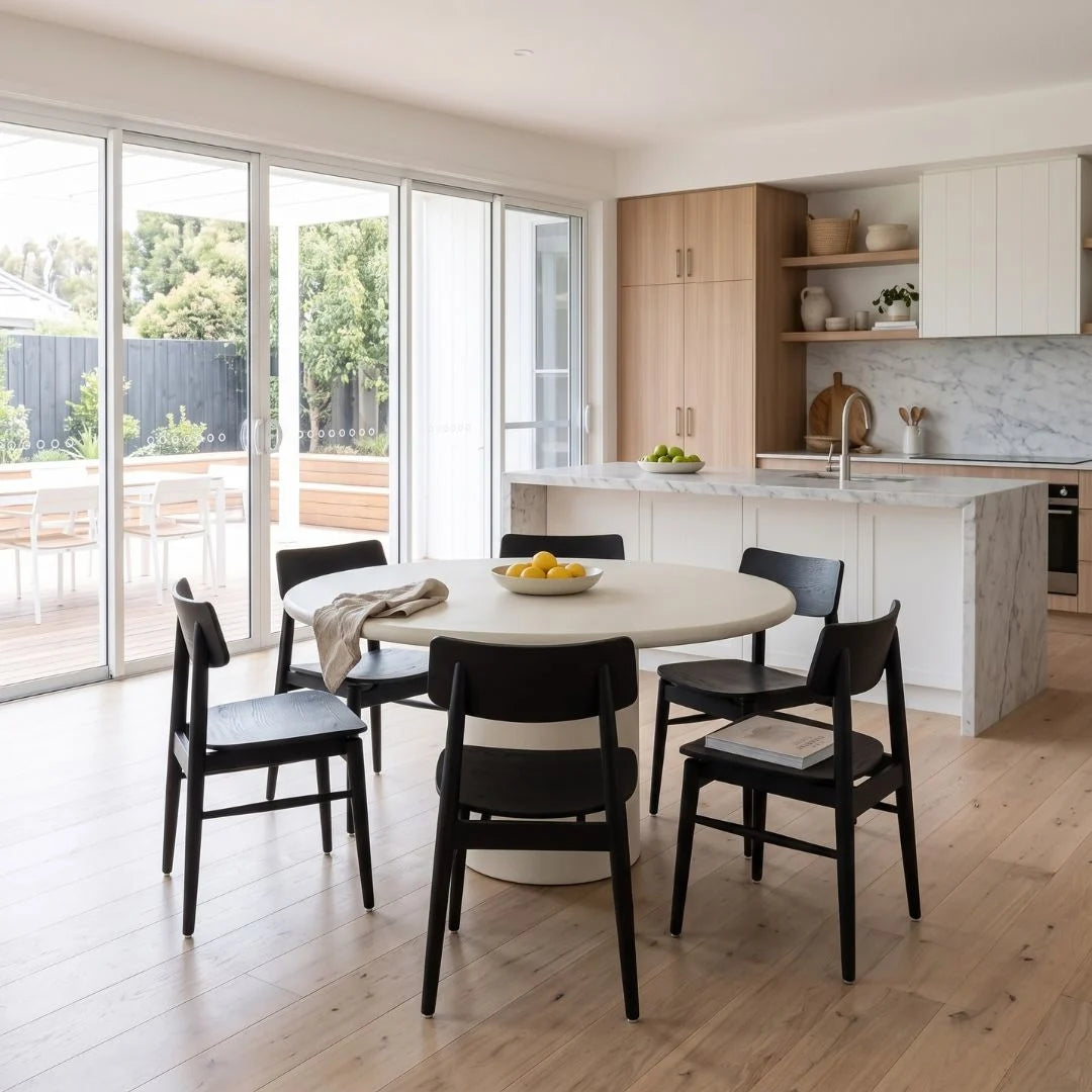 A modern dining room setup featuring a round white table with black chairs, set in a bright, contemporary kitchen with light wood cabinets and marble countertops.