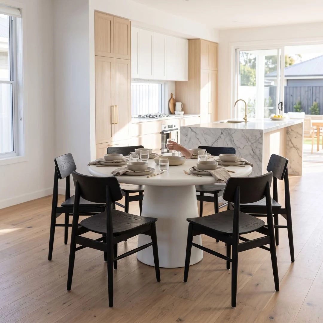 A dining room scene with a round white table surrounded by six black wooden chairs, set with white plates and glasses. The room features light wood flooring, white walls, and a modern kitchen in the background with white and light wood cabinets and a marble island.