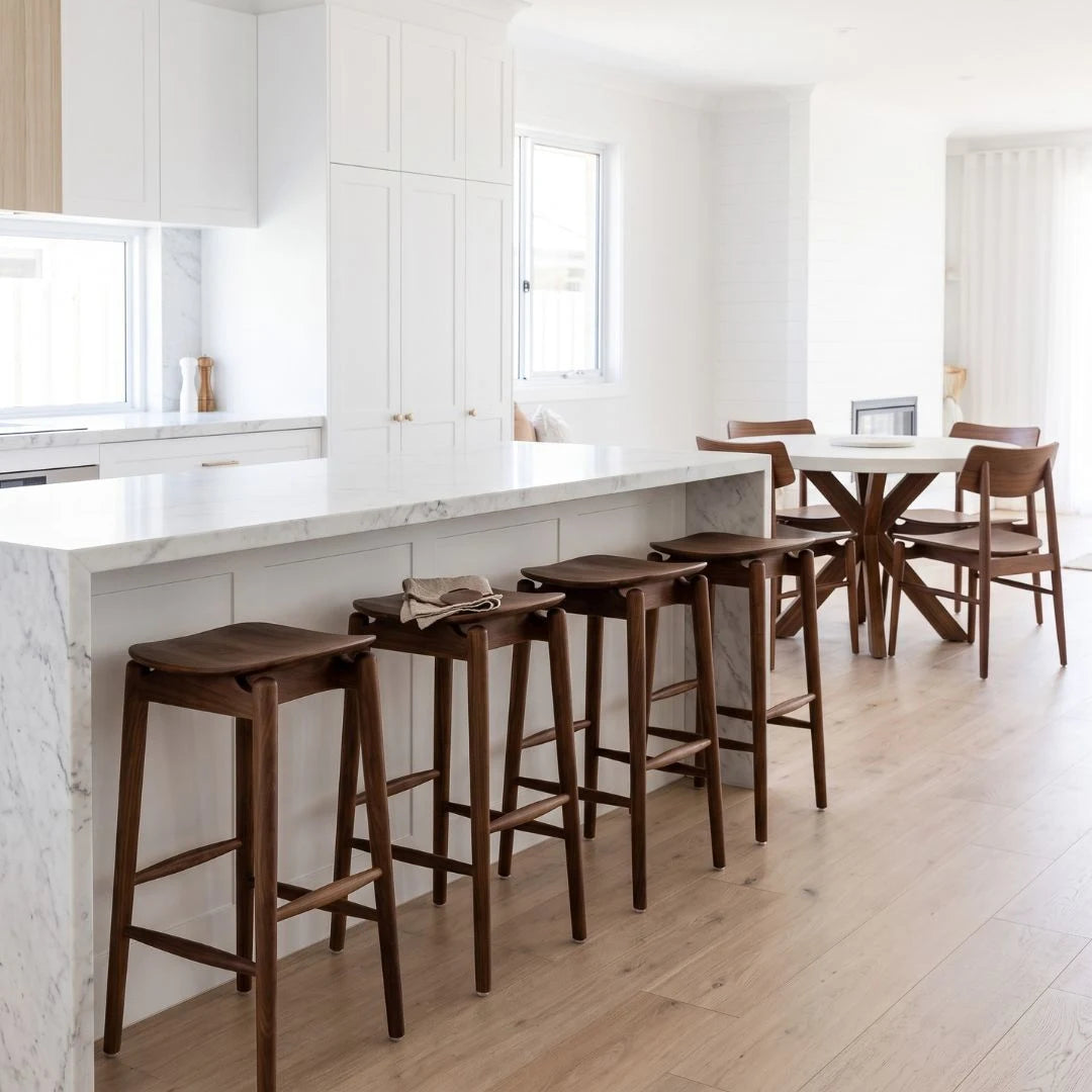Four dark wood bar stools with white marble-topped seats arranged along a white kitchen island in a bright, modern kitchen with light wood floors.