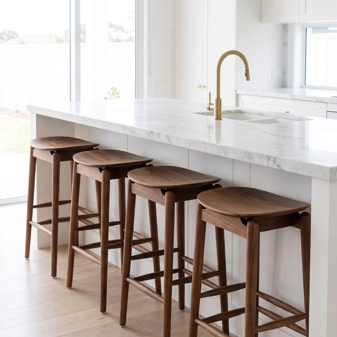 Four wooden bar stools with dark brown seats and legs arranged around a white kitchen island with a marble countertop and a gold faucet.