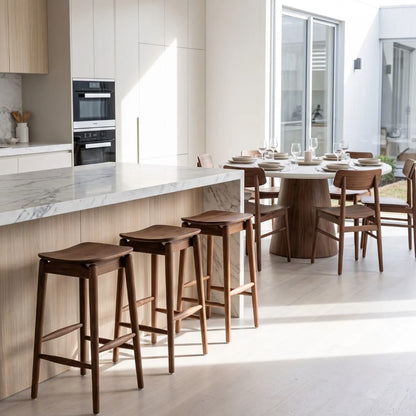 A modern kitchen and dining area with four dark wood bar stools positioned along a marble-topped island, next to a round dining table and chairs.
