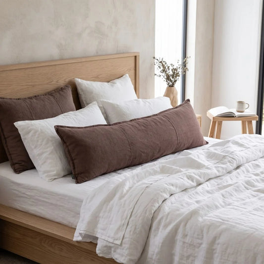 A styled bedroom scene with a bed dressed in white bedding and two long brown lumbar pillows, set against a light wood headboard and a softly lit neutral interior.