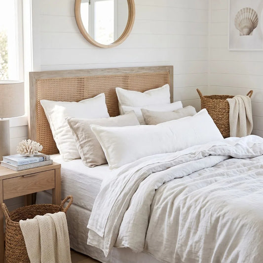 A neatly made bed with white and beige linens, a woven cane headboard, and a softly lit bedroom with a nightstand, basket, and shell artwork on the wall.