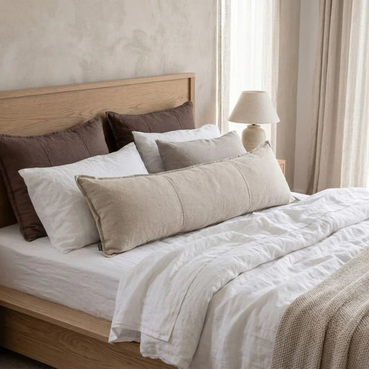 A neatly made bed with white linens, a light wood headboard and footboard, and a mix of neutral pillows including a prominent beige lumbar pillow, styled in a softly lit bedroom.