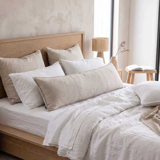 A neatly made bed with white and beige linens, multiple pillows, a light wood headboard, and a matching wooden footboard in a softly lit bedroom.
