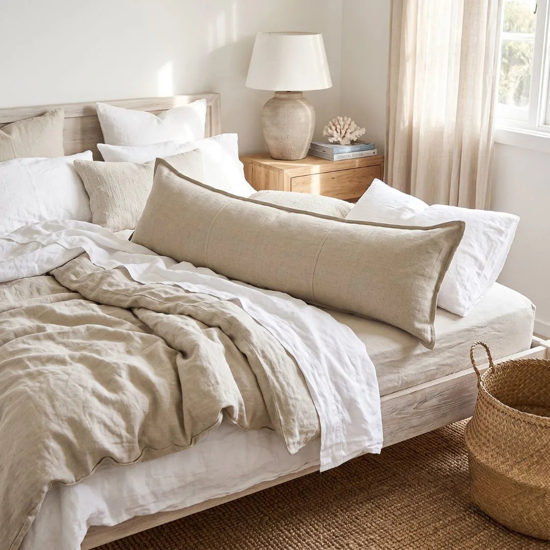 A long, rectangular linen lumbar pillow in a light beige color rests on a bed styled with white linens, a wooden headboard, a bedside table with a lamp, and a woven basket on the floor.