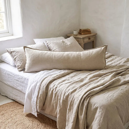 A beige linen lumbar pillow resting on a bed with white linens and a matching beige throw blanket in a softly lit, rustic bedroom.